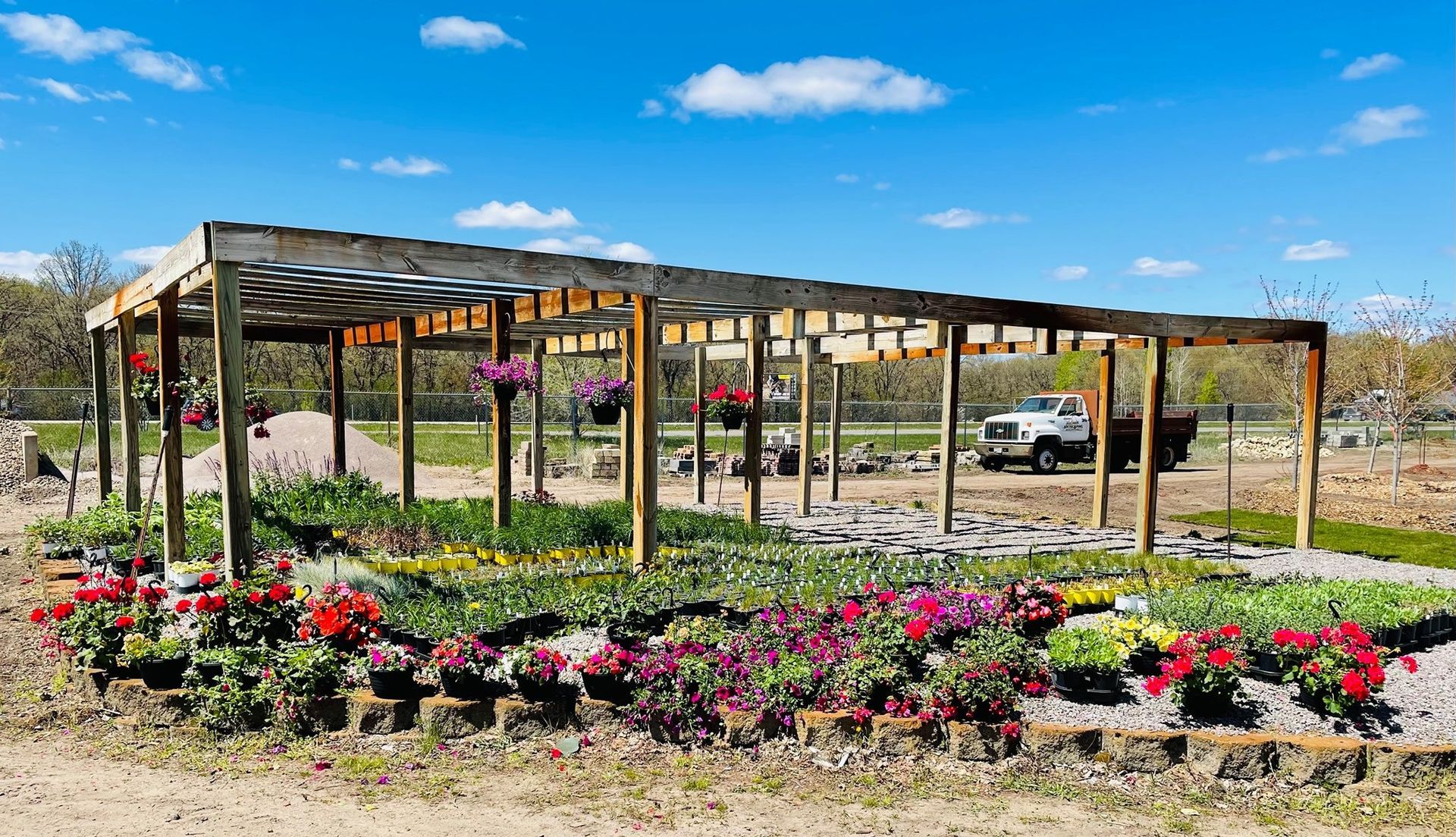 Colorful flower beds under a wooden pergola in a sunny outdoor nursery with a truck in the background