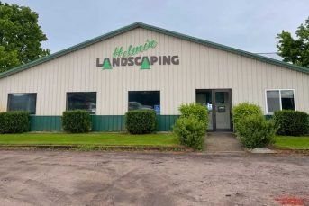 Front view of a beige landscaping business building with green trim and sign, bordered by shrubs and gravel lot