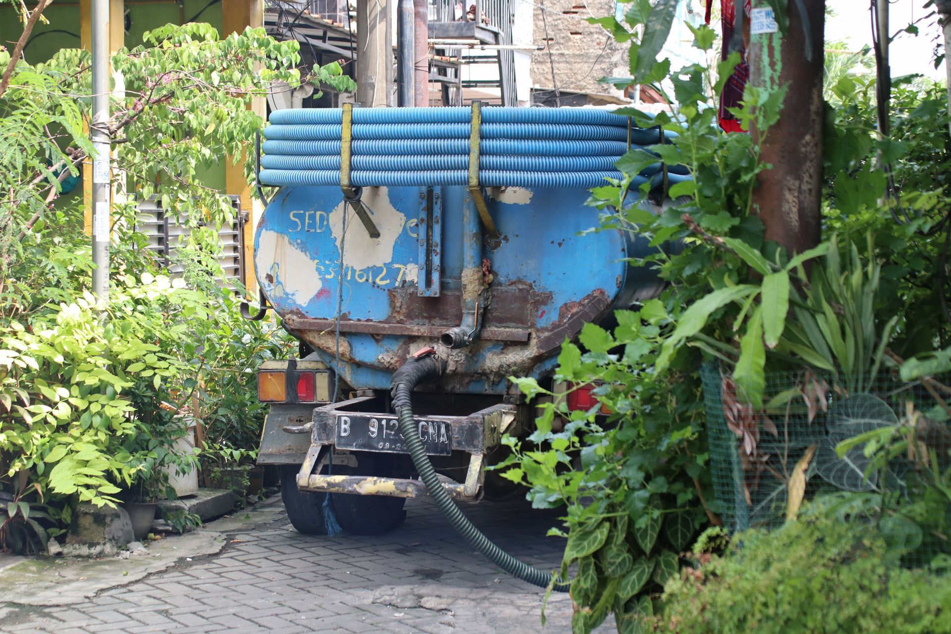 Blue water truck parked on a narrow road surrounded by greenery.