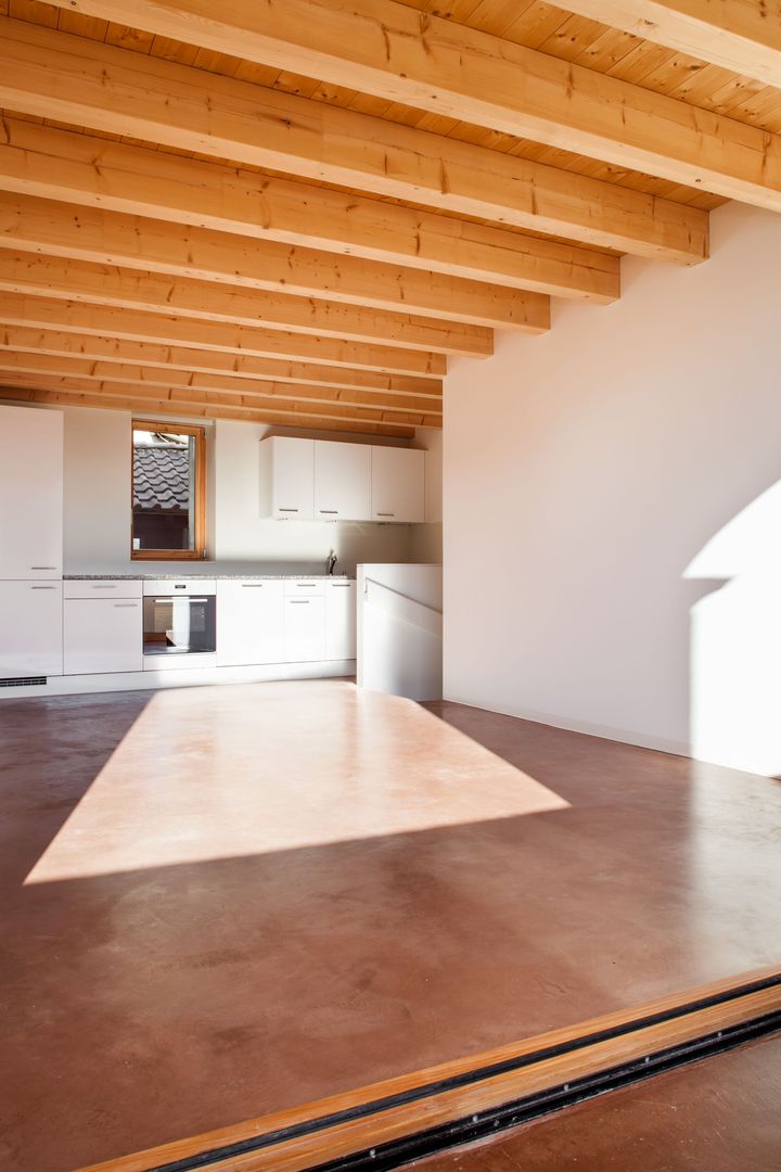 Empty modern room with exposed wood beams, white walls, and a small kitchen area in the back.