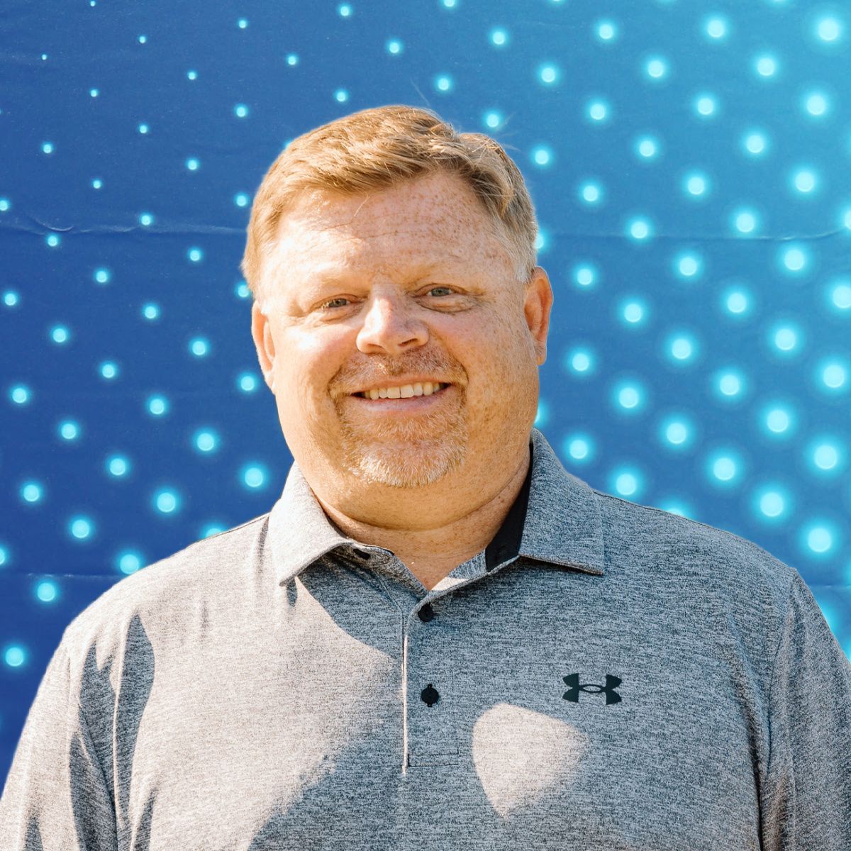 Smiling man in a gray polo shirt standing before a blue dotted backdrop