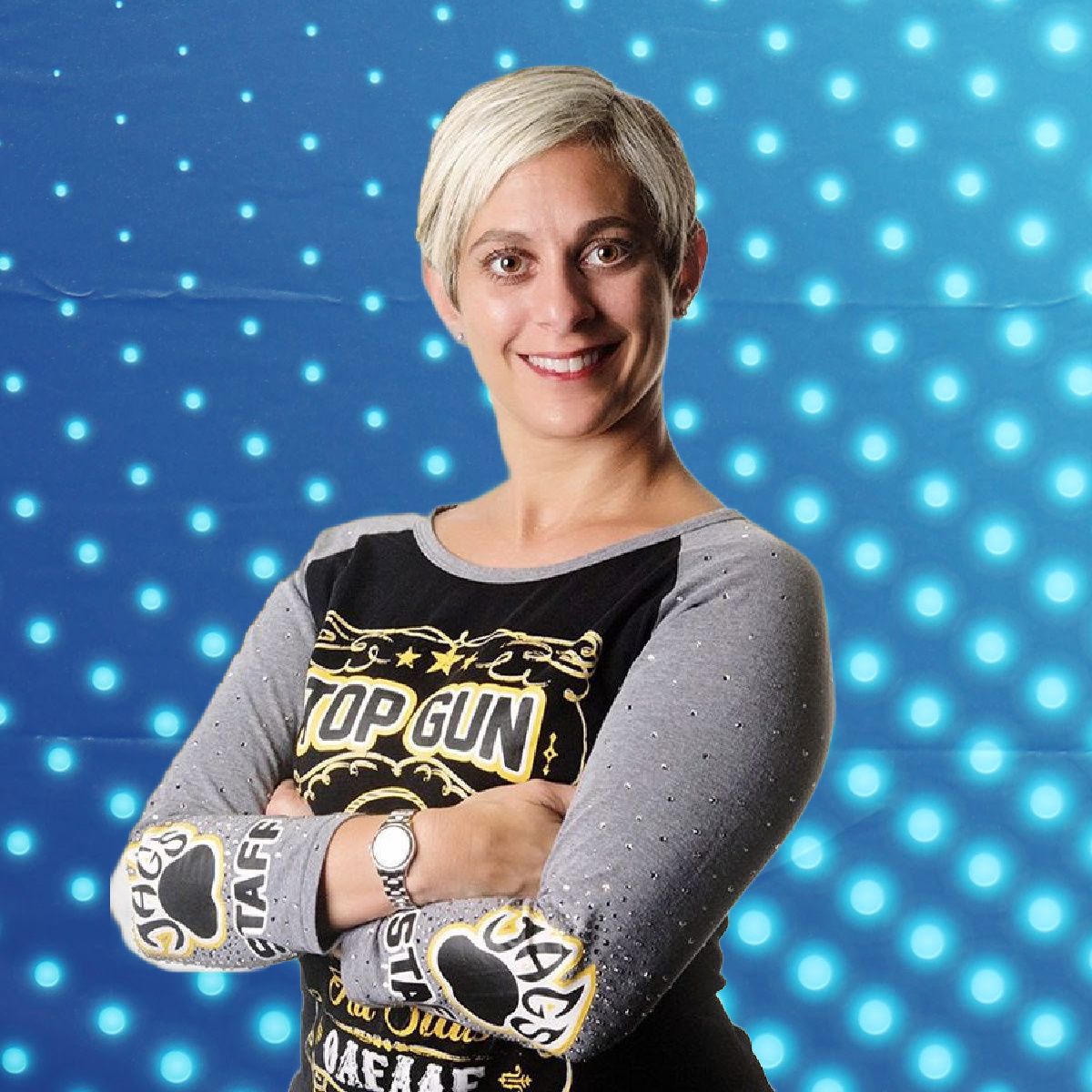 Smiling person with short blonde hair and crossed arms, wearing a Top Gun shirt against a blue dotted background