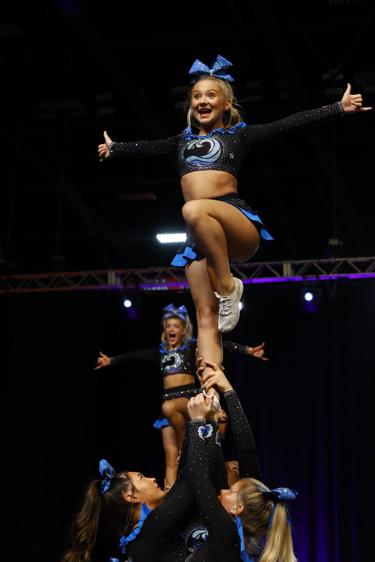 Cheerleader stunt in progress: smiling flyer held high with arms outstretched under stage lights