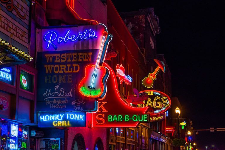 Neon signs on a lively New Orleans street, including Roberto’s, Western World, and a guitar sign.