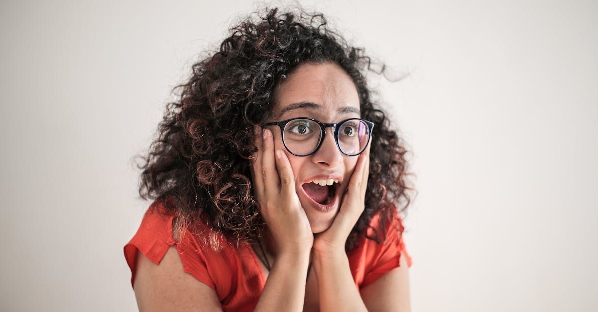 Shocked person with curly hair and glasses, hands on cheeks, against a plain background