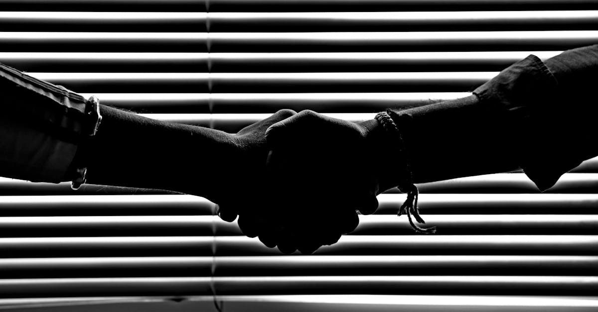 Two hands clasped in a handshake silhouette against horizontal blinds.