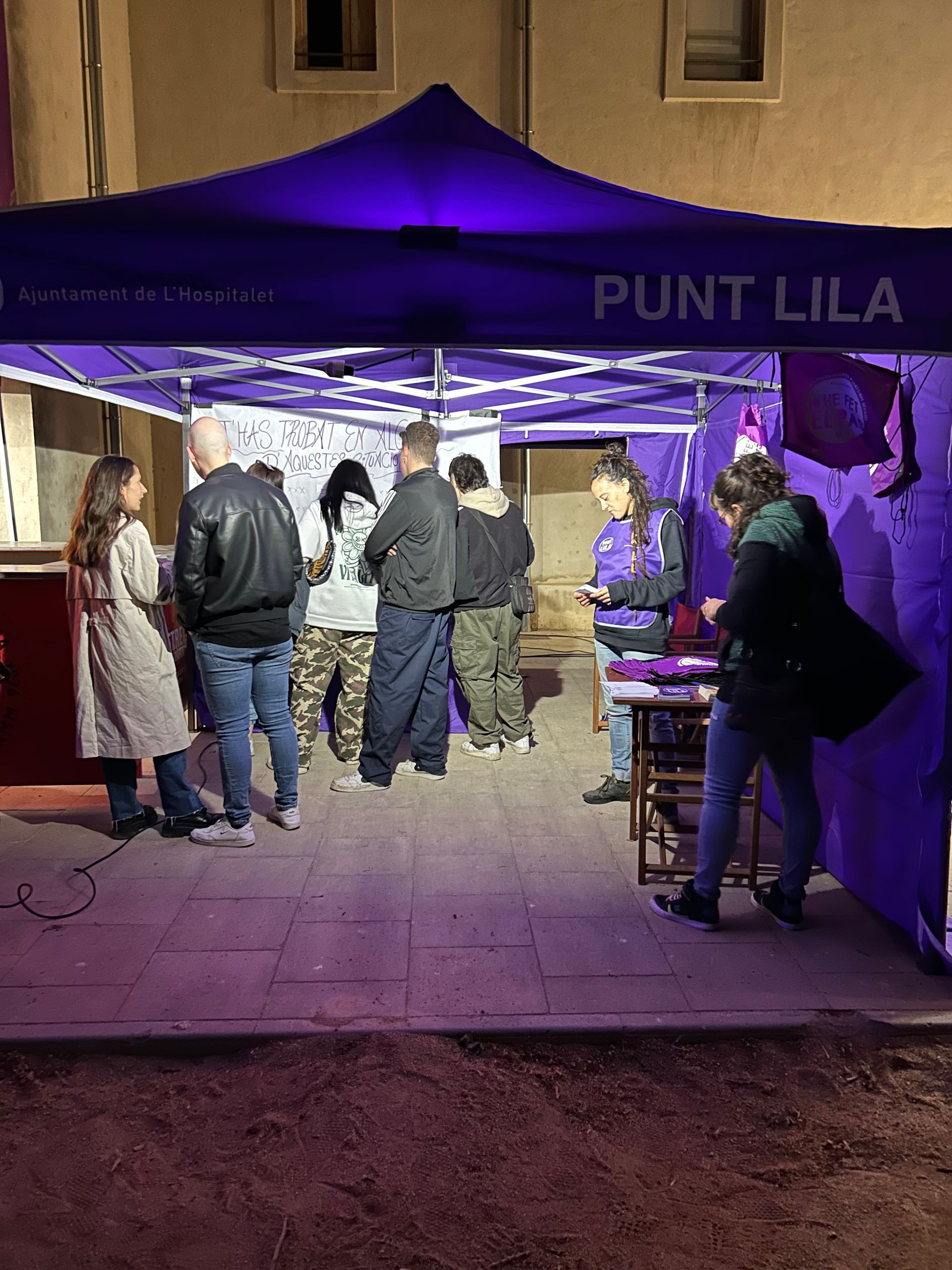 La gent es reuneix en una tenda lila de Punto Lila, possiblement una estació de suport. Ambient nocturn, il·luminat.