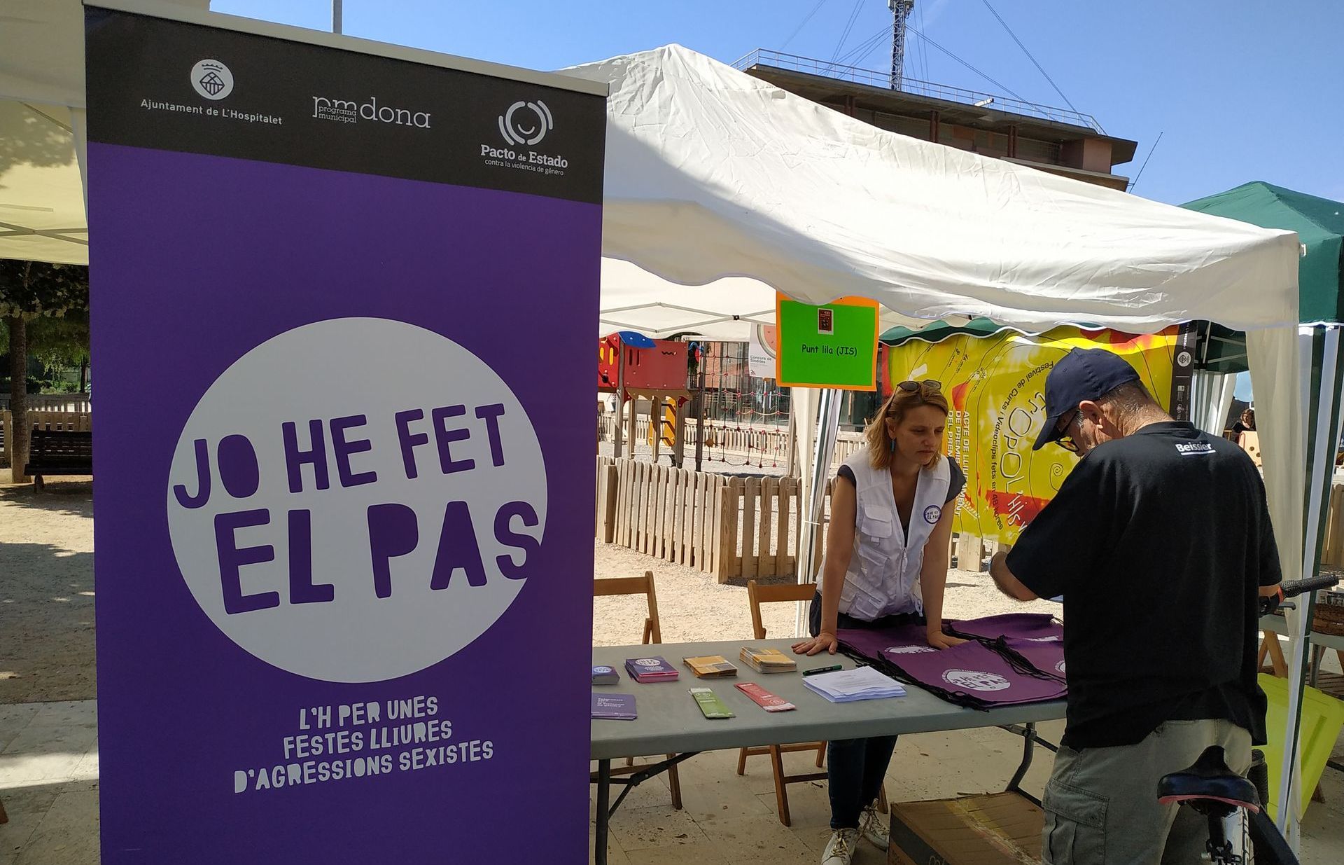 Una dona i un home en una parada amb pancartes morades que promouen una iniciativa de salut pública en un mercat a l'aire lliure.
