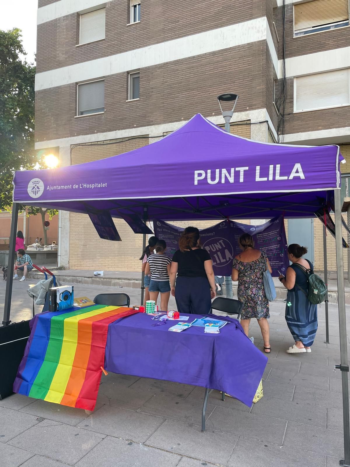 Carpa lila de Punto Lila amb una bandera arc de Sant Martí, dones a una taula i edifici de maó al fons.