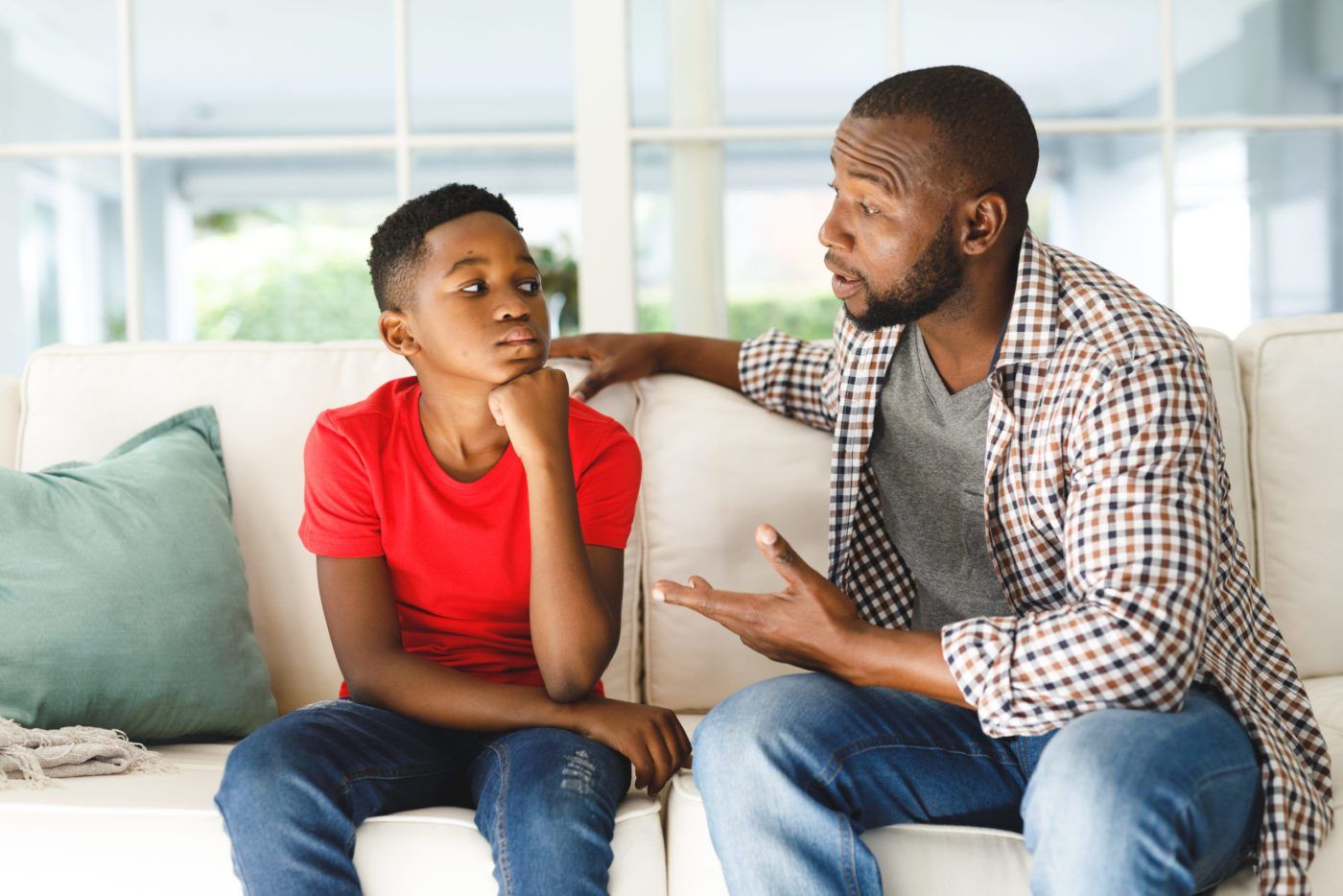 Two people talking on a couch in a bright living room, one gesturing while the other listens attentively