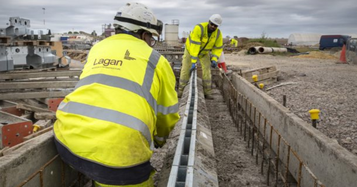 Construction workers in yellow vests install a metal drain in a concrete trench at an outdoor construction site.