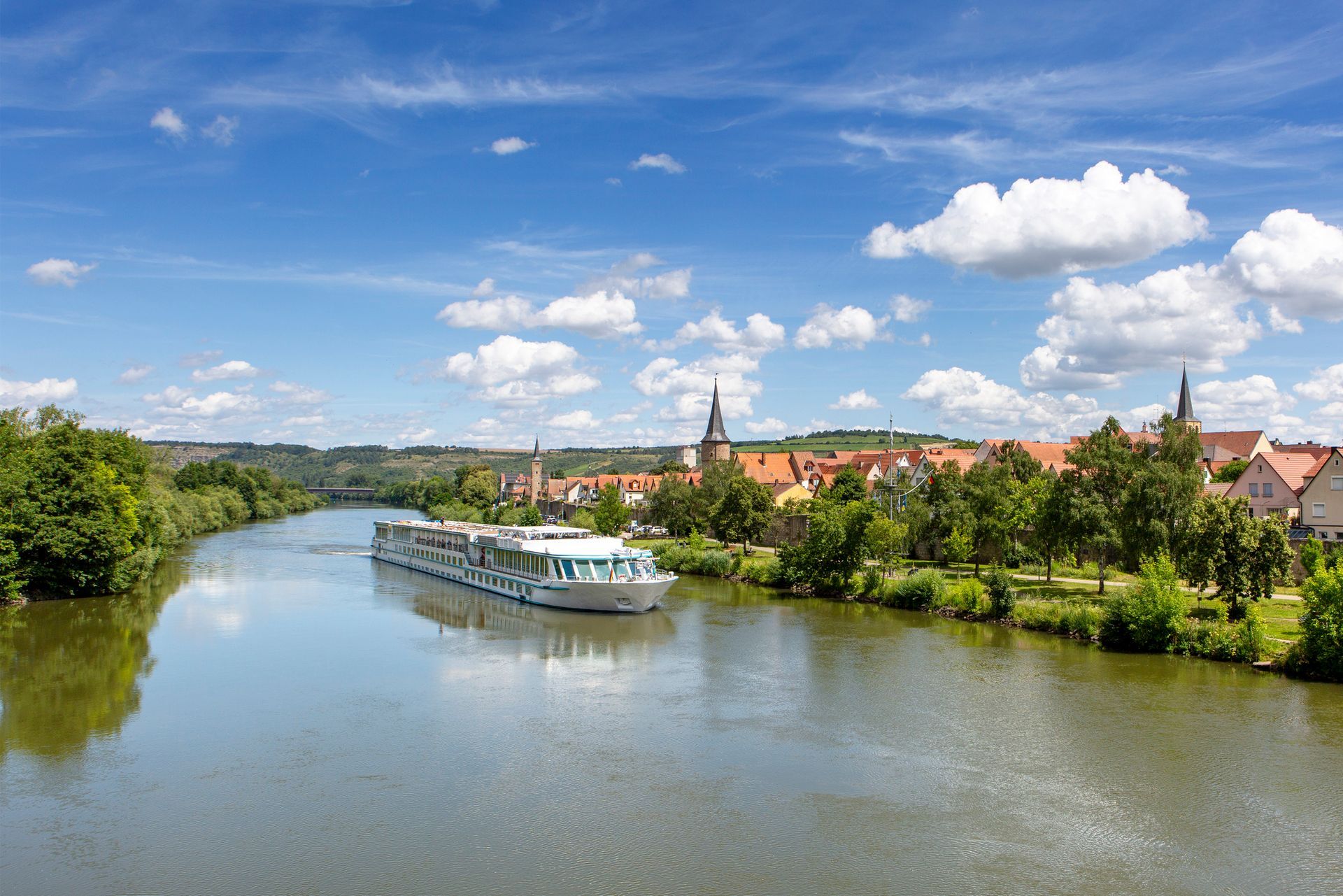 River cruise ship on a calm river with a town on the shore under a blue sky.
