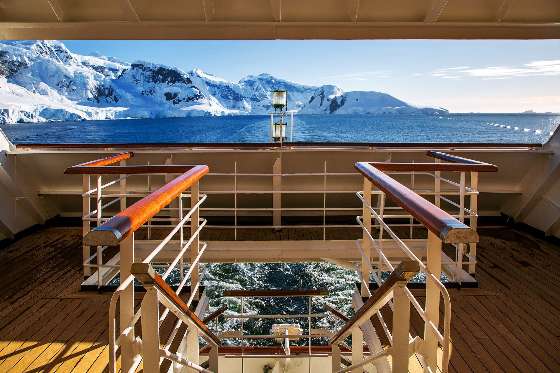 View from cruise ship deck to snow-covered mountains over blue water. Wooden railings and deck.