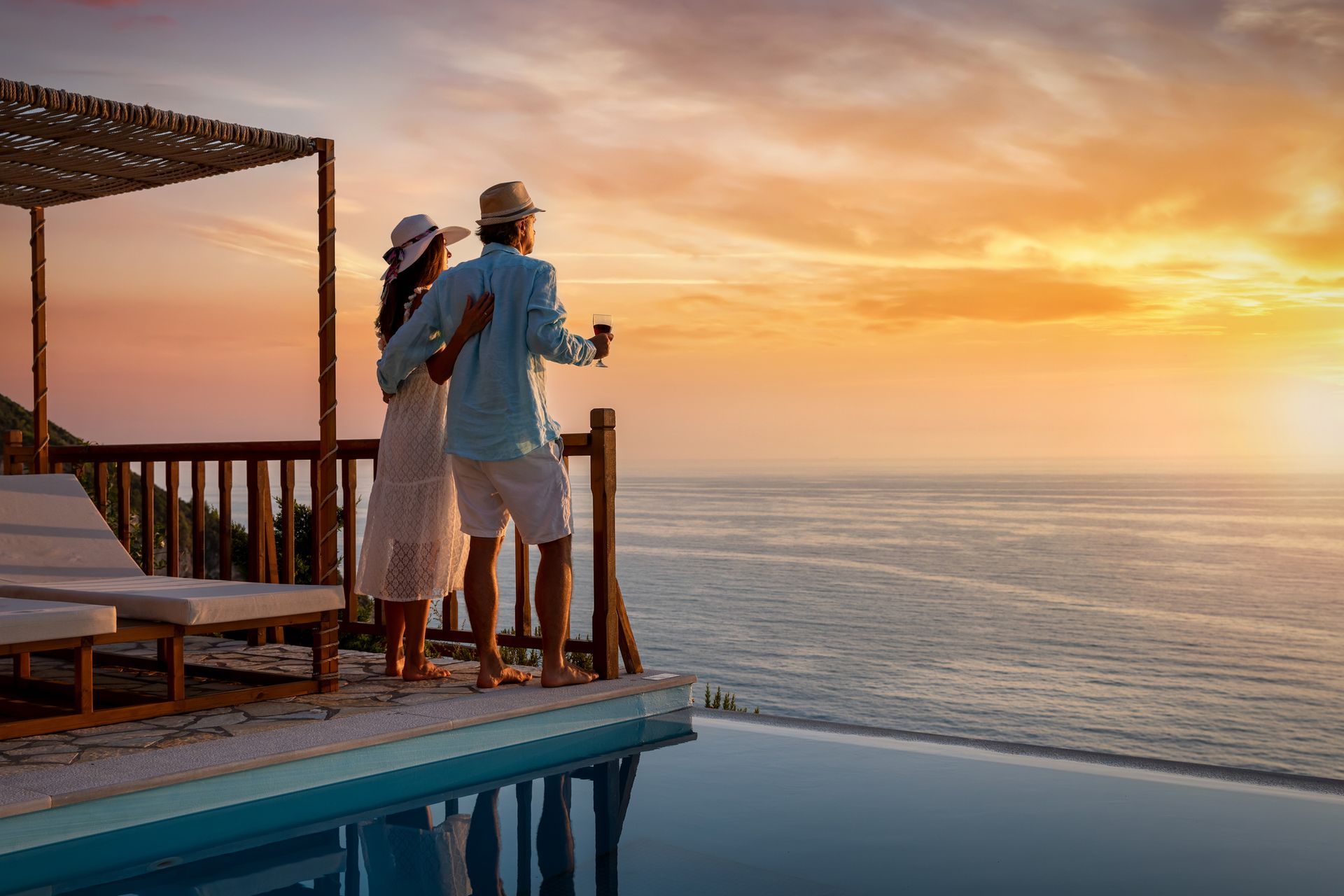 Couple on balcony overlooking ocean at sunset, holding drinks; pool in foreground.