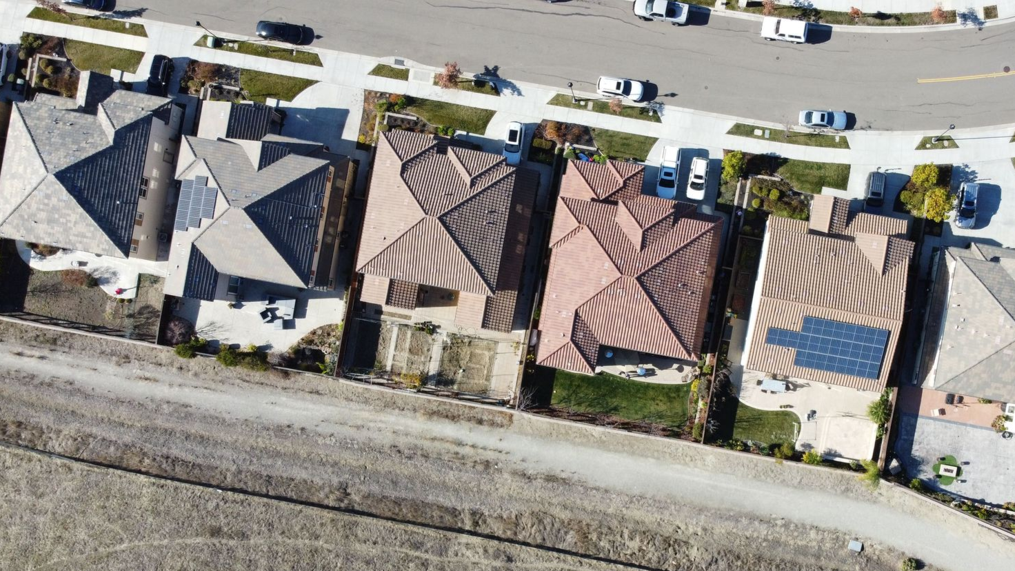 Aerial view of four suburban homes with tiled roofs, parked cars, and a road along the top and railroad tracks below.