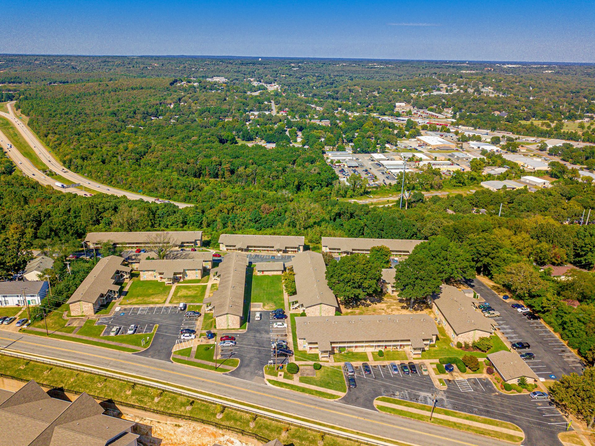 Aerial view of apartment complex with parking, road, and surrounding forest on a sunny day.