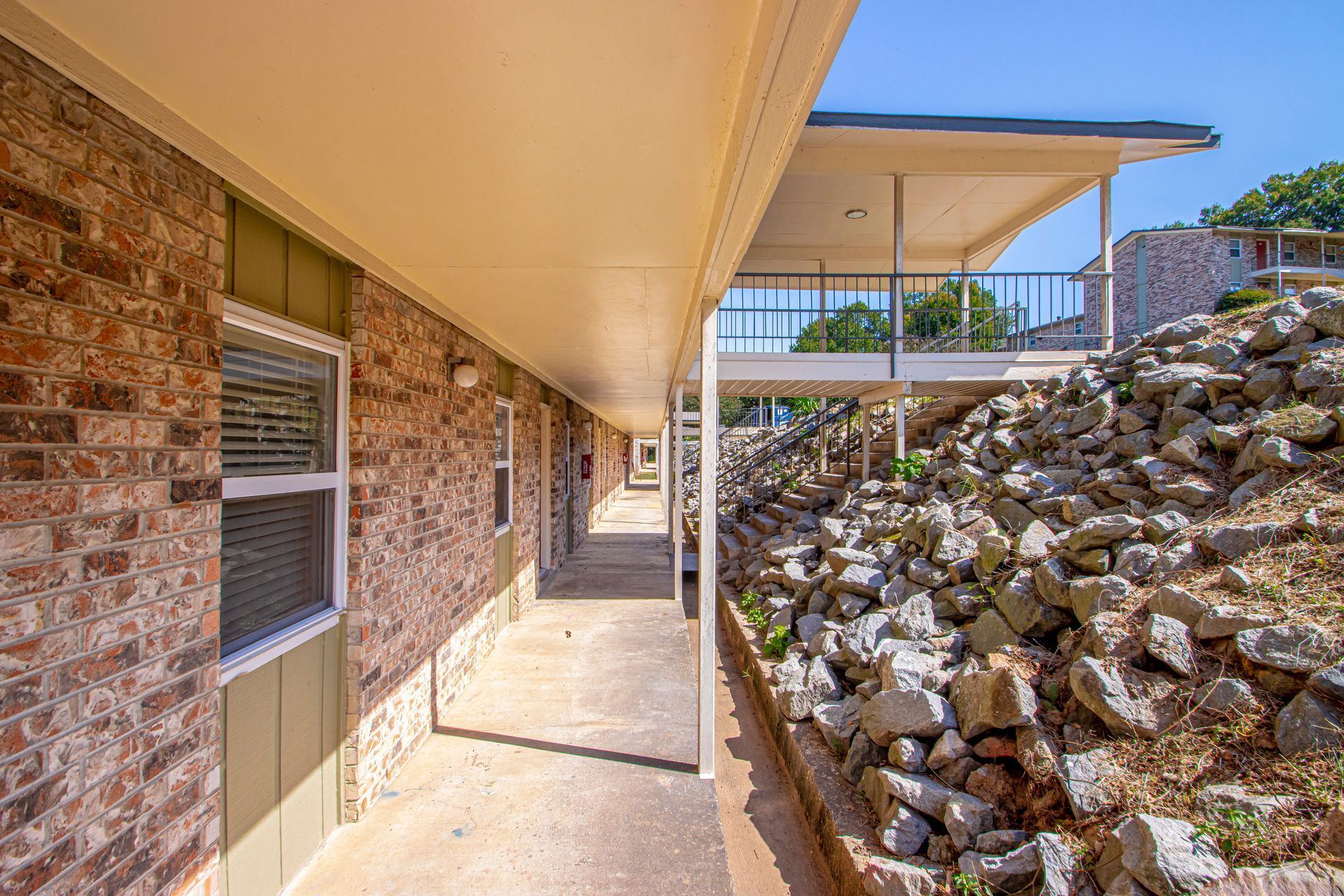 Walkway alongside a brick building with a covered walkway. A rocky hillside is on the right.