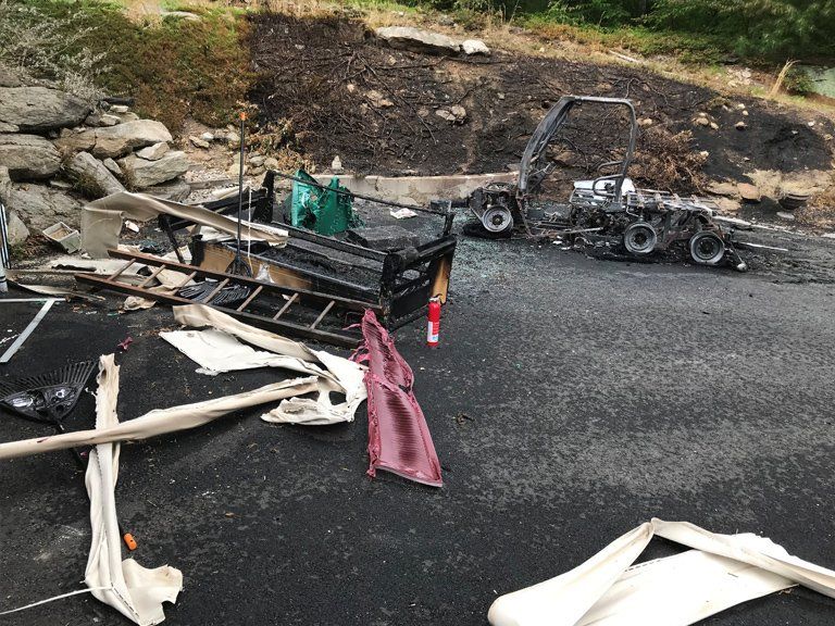Charred remains of a vehicle and debris scattered on asphalt, with burnt hillside in the background.