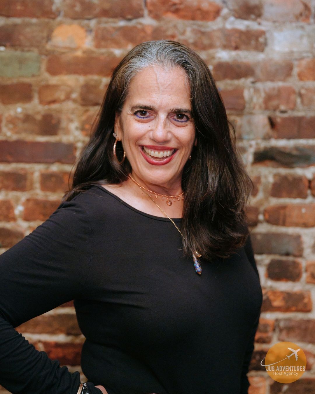 Woman smiles in front of a brick wall, wearing a black long-sleeved shirt and gold jewelry.