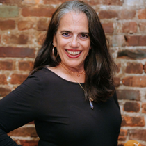 Woman smiles in front of a brick wall, wearing a black top and jewelry.