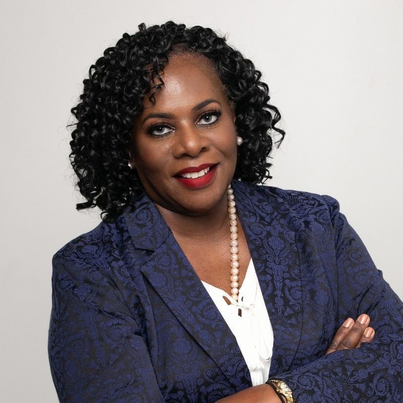 Woman with dark curly hair in blue patterned blazer, arms crossed, smiling.