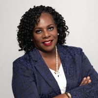 Woman with dark curly hair in blue patterned blazer, arms crossed, smiling.