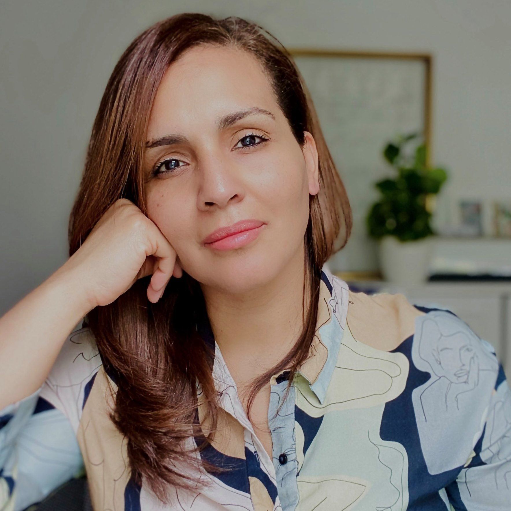 Woman with brown hair resting her hand on her cheek, wearing a patterned shirt, looking at the camera.