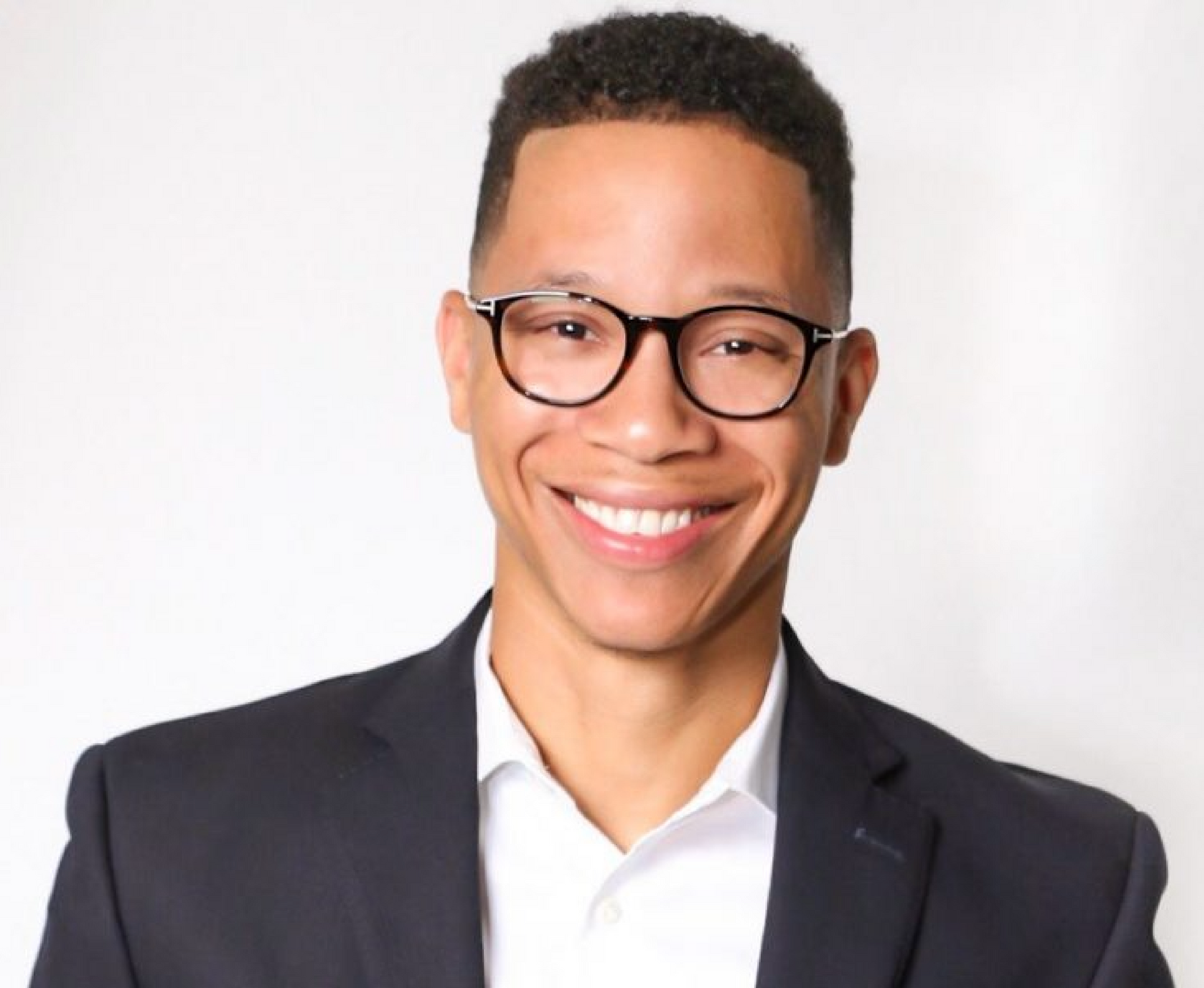 Man with glasses and a suit smiling at the camera against a white background.