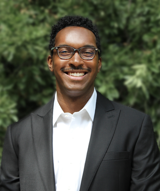 A Beltino Goncalves in a black blazer and white shirt smiling outdoors in front of a blurry green background.