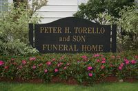 Sign for Peter H. Torello and Son Funeral Home, black with gold lettering, surrounded by flowers and greenery.
