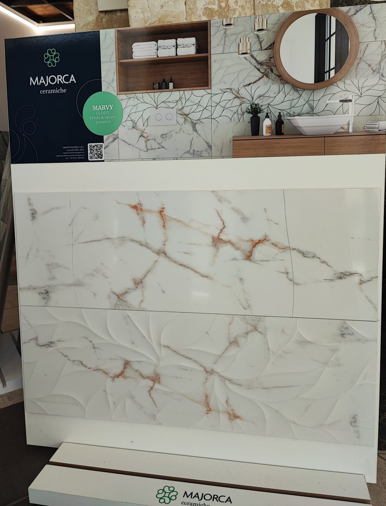 Esposizione di piastrelle effetto marmo con venature dorate, dietro una cabina armadio con specchio, lavandino e mensole.