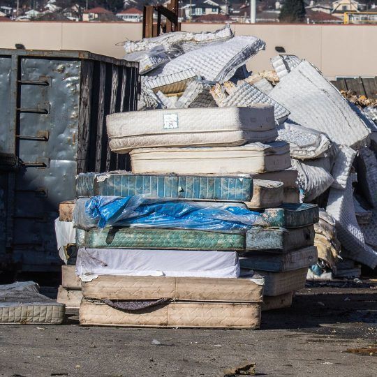 A pile of mattresses sits in front of a dumpster