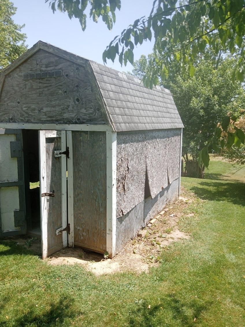 A shed with a roof and a door is in the middle of a grassy field.