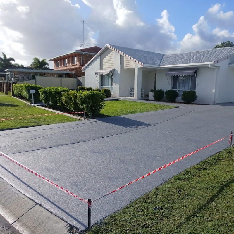 White house with a blue roof and freshly paved driveway, bordered by red caution tape on a sunny day — Troys Concrete Overlays in Hillcrest, QLD