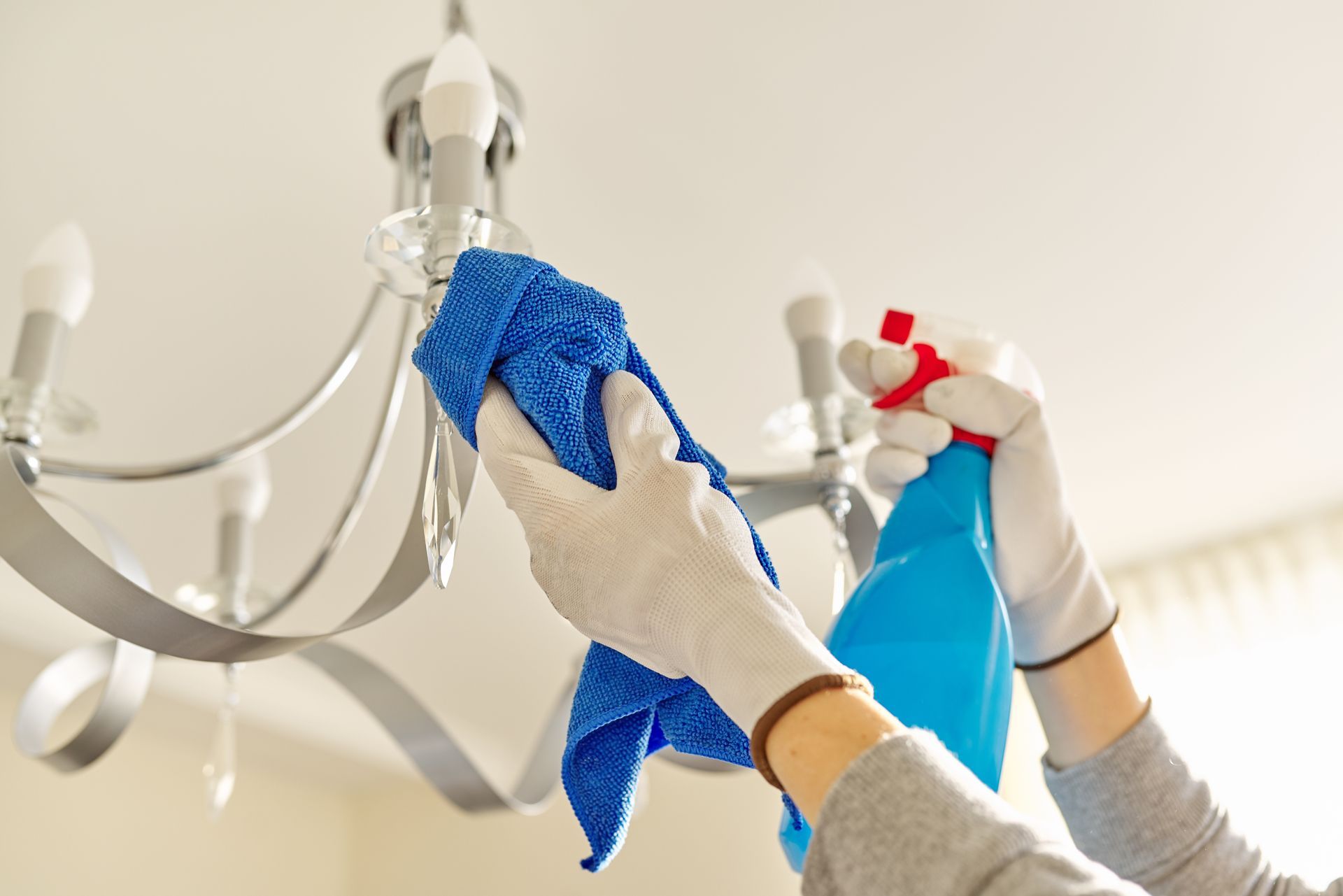 Hands wiping a ceiling light fixture with blue cloths and a spray bottle in a bright room