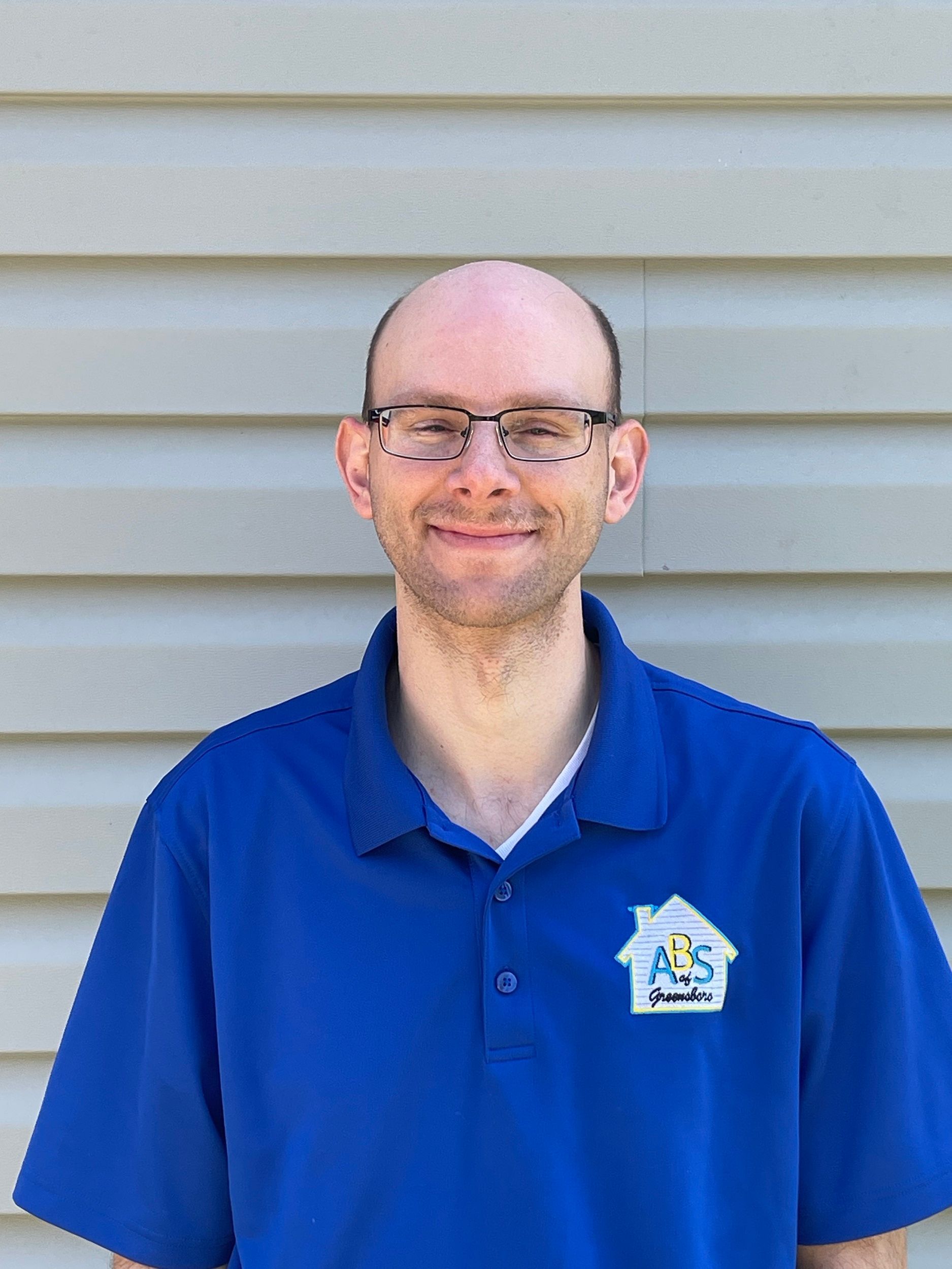 Man in blue polo shirt and glasses standing against a beige siding wall, smiling