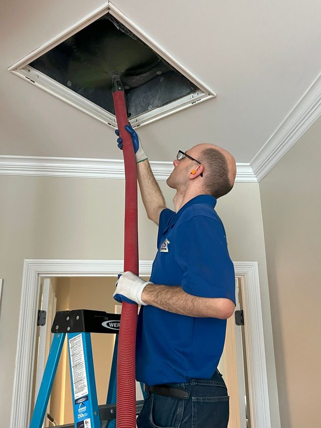 Man reaching into a ceiling vent with a red vacuum hose during home maintenance