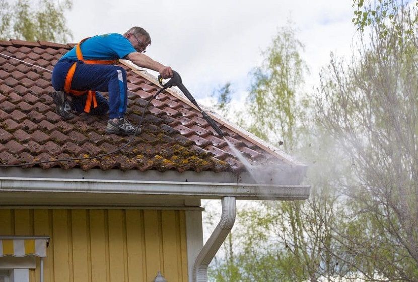 Worker on a roof using a pressure washer to clean moss from red shingles
