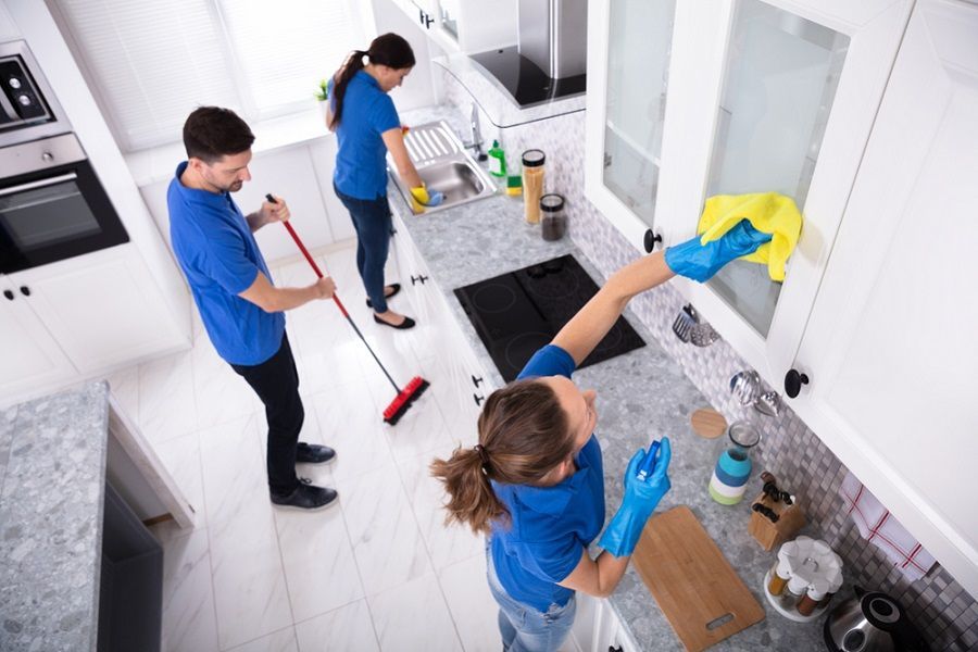 Three people cleaning a bright kitchen with mops, cloths, and spray bottles.
