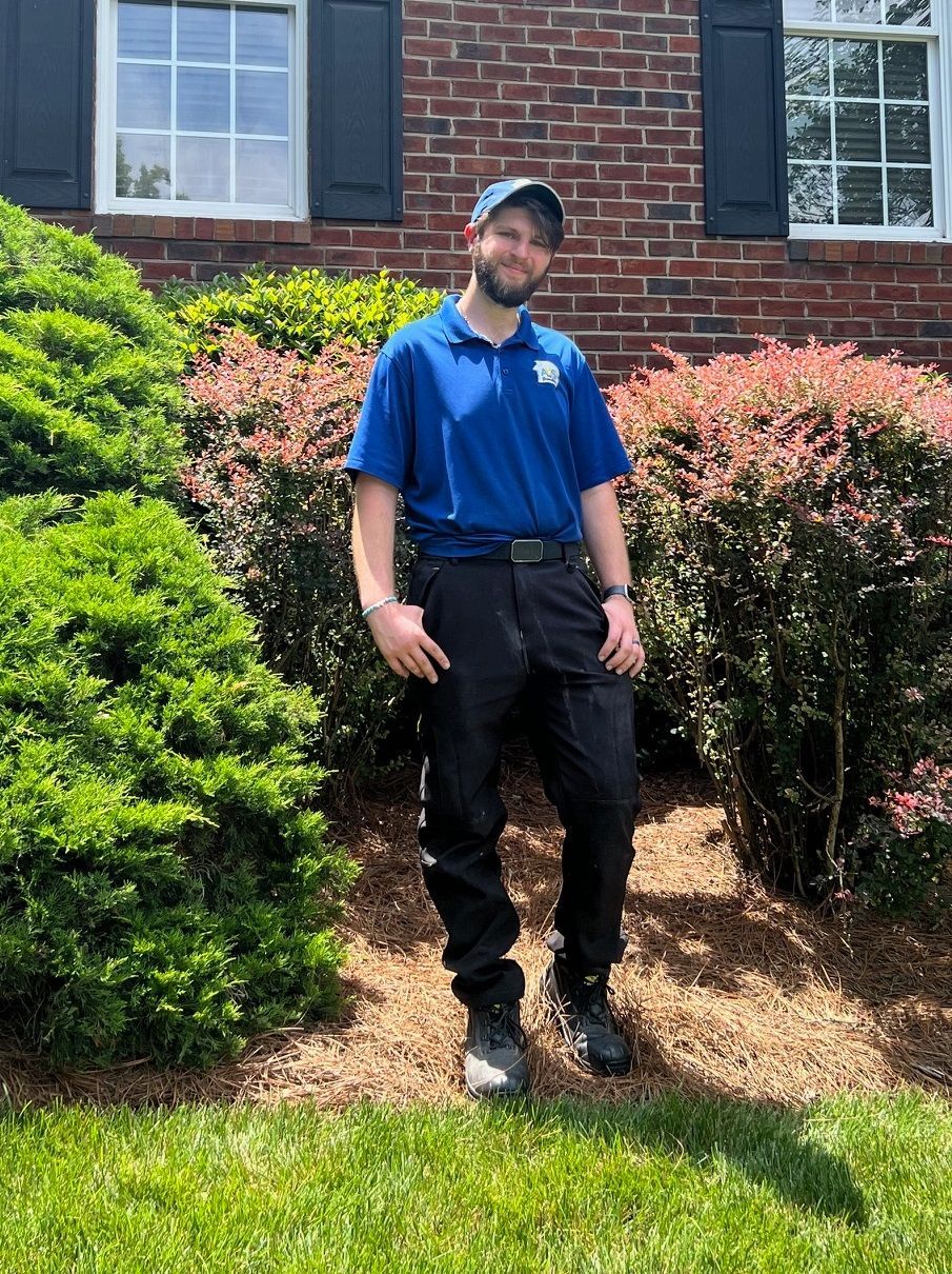 Man in blue shirt and black pants standing in a garden in front of a brick house.