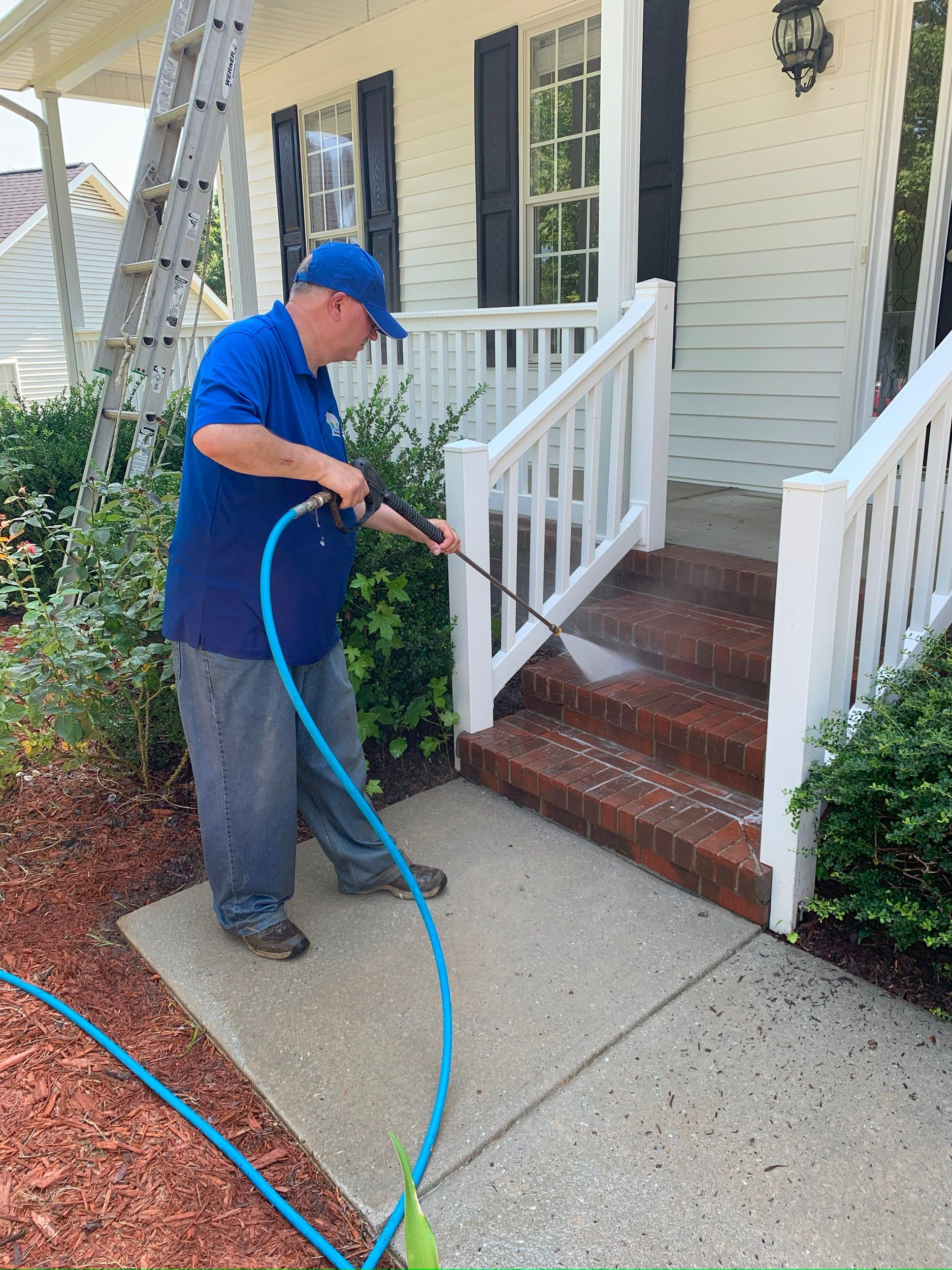 Person pressure washing brick steps and a front entry walkway with a blue hose.