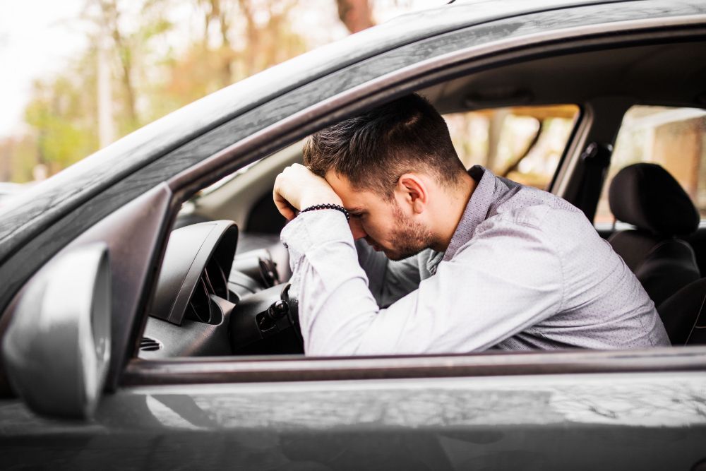 Man in car, head in hands, appears stressed. Grey car, daytime setting.
