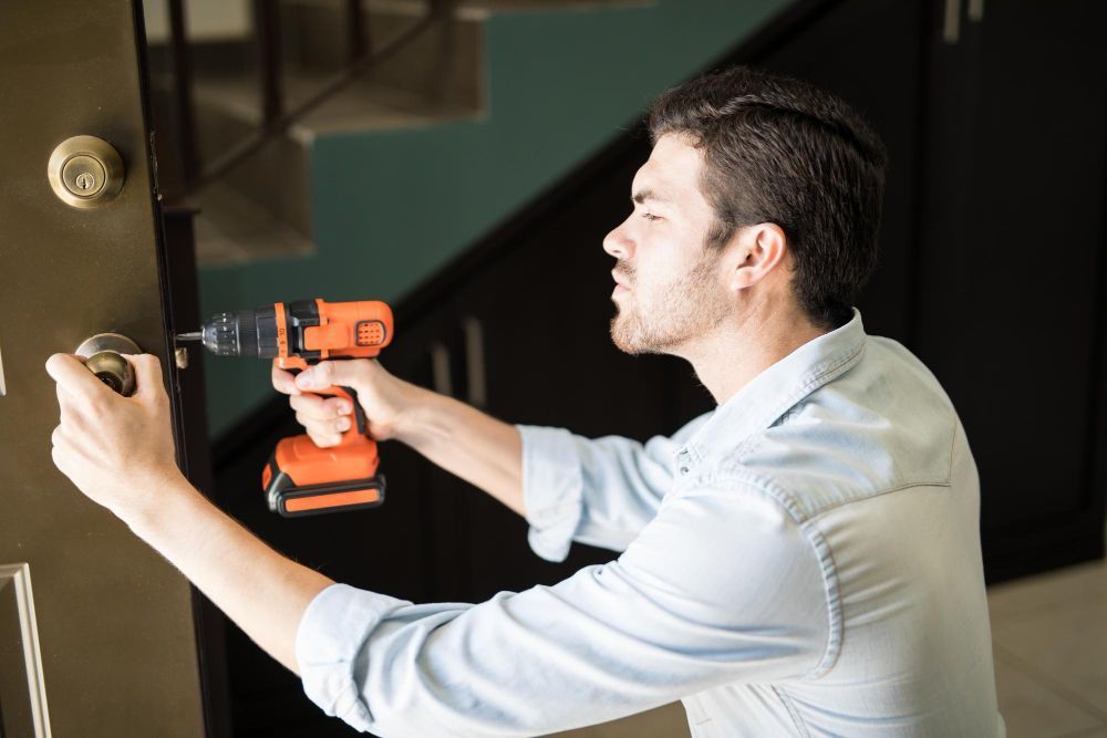 Man using an orange drill to install a doorknob on a brown door inside a house.