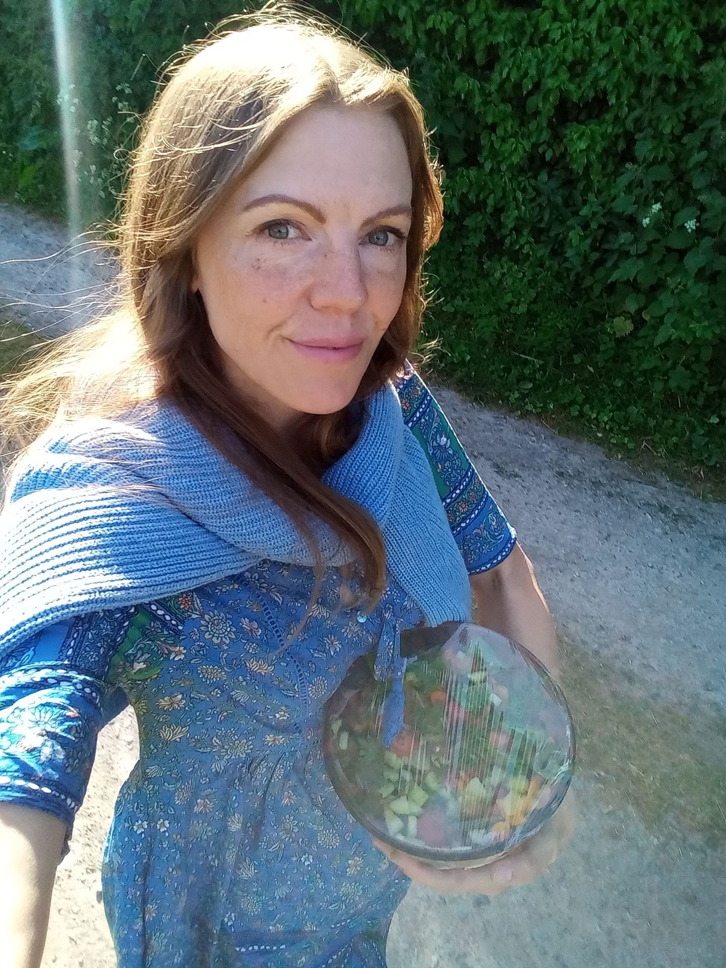 Person in a blue patterned top taking a selfie outdoors beside green hedges, holding a bowl of colorful plants.