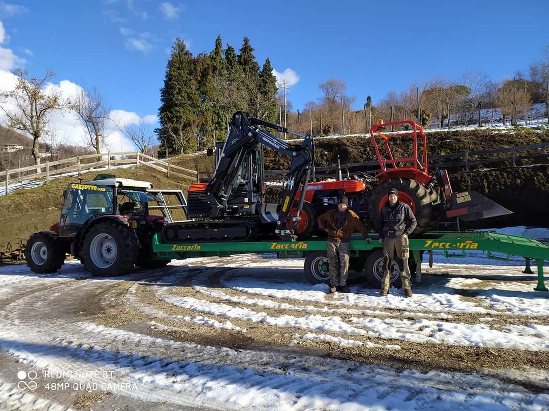Due persone sono in piedi vicino a un rimorchio verde che trasporta trattori e un escavatore in un campo innevato in una giornata di sole.