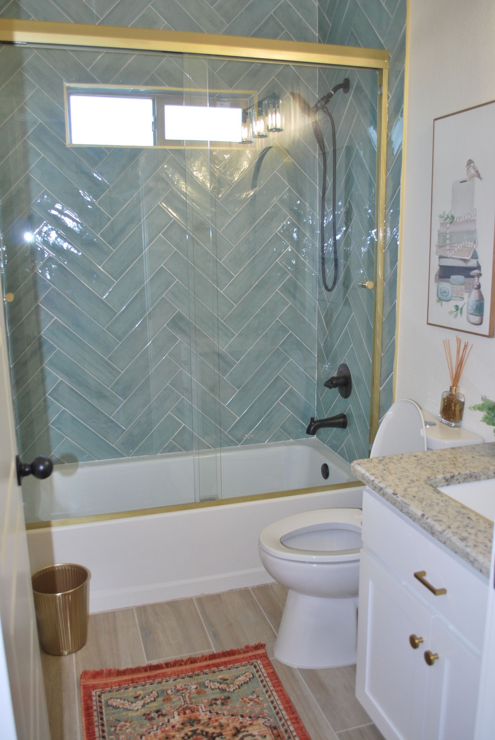 Bathroom with teal herringbone tile, gold accents, white vanity, toilet, and rug.