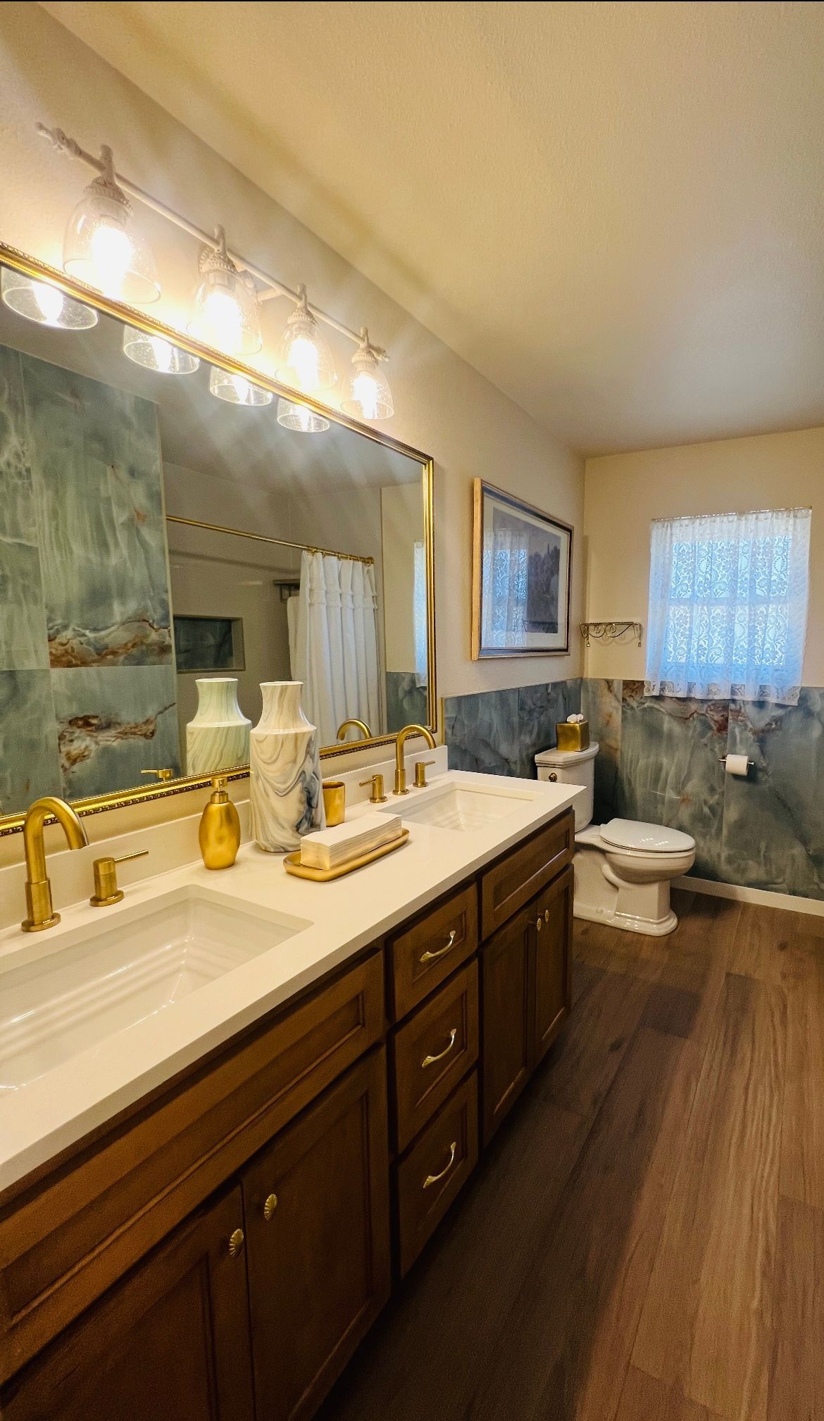 Bathroom with double sinks, wooden vanity, gold fixtures, and blue-green tiled walls.