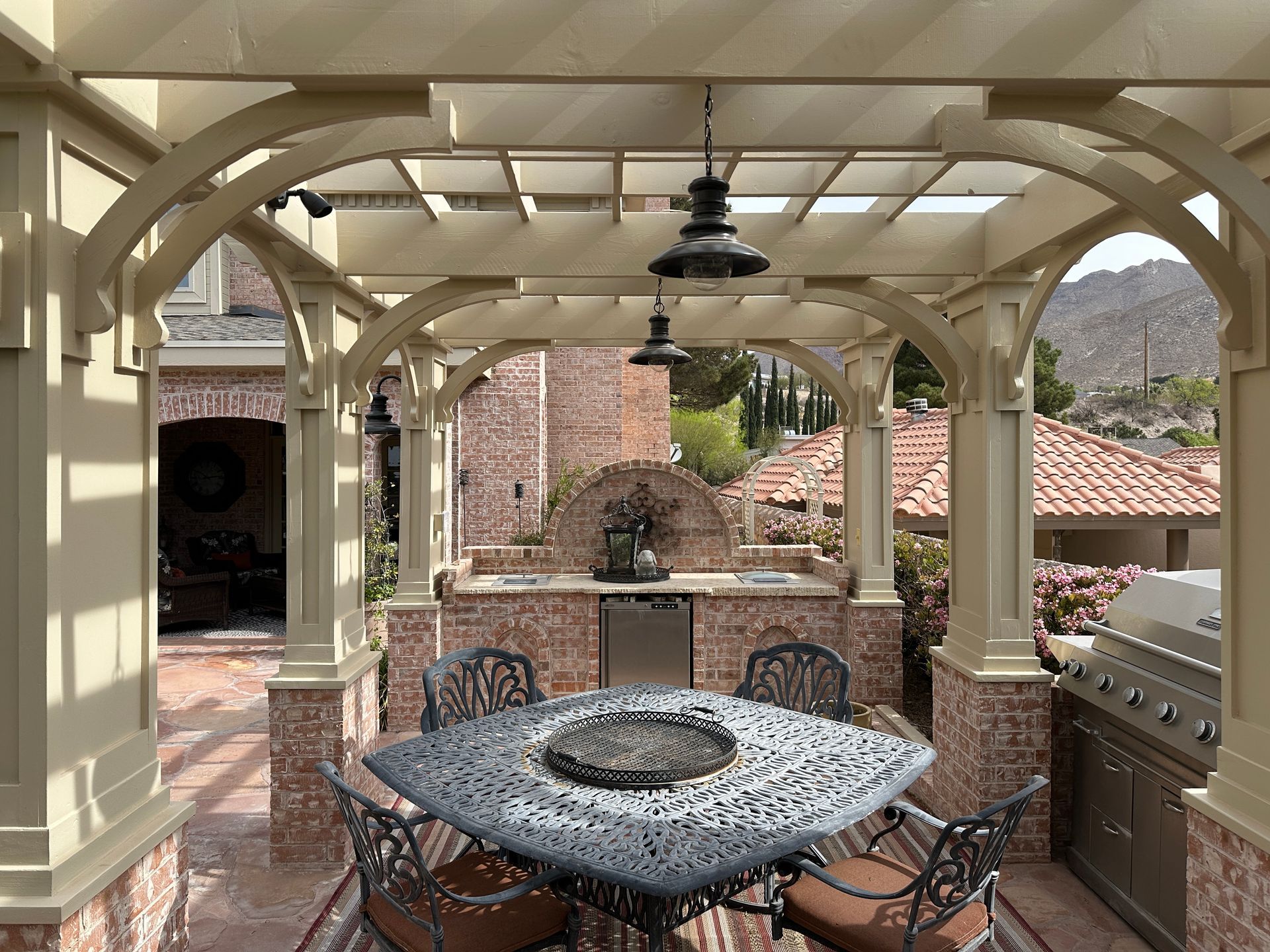 Outdoor kitchen with stone and brick, under a wooden pergola, dining set, and grill.