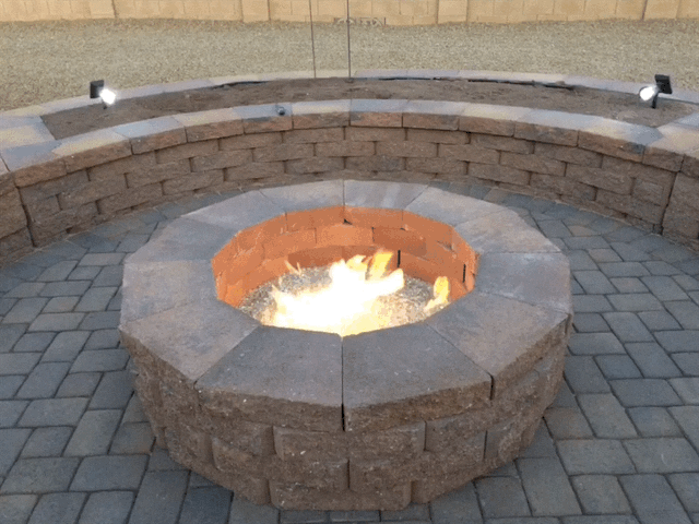 Fire pit with flames surrounded by brick seating on a paved patio, lit by small lights.