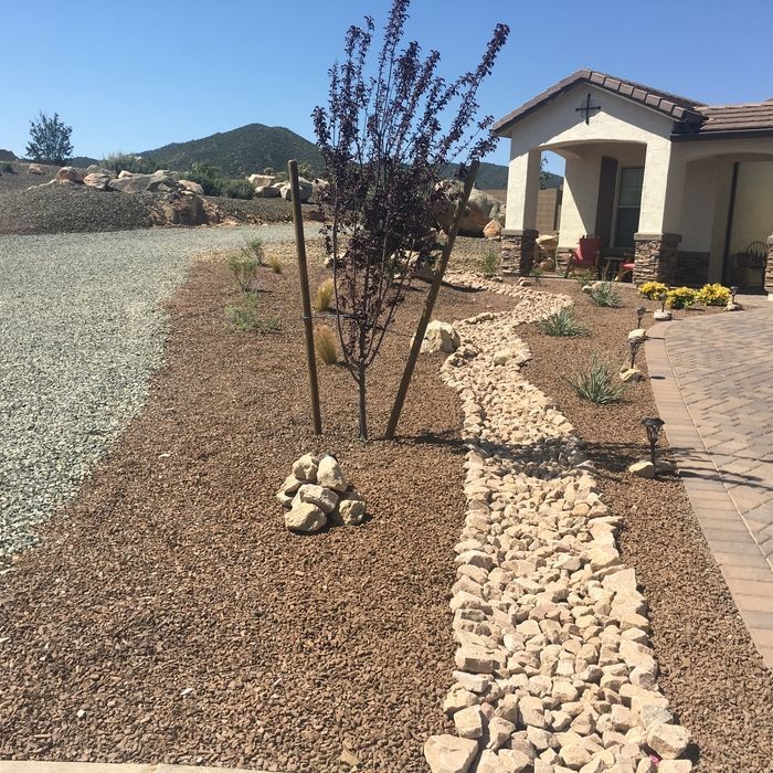 Desert front yard with gravel, rock border, small tree, and a stucco house beside a paved walkway